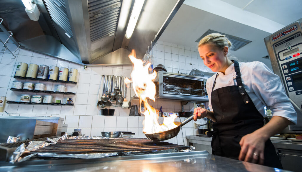 Restaurantküche, in der eine Köchin gerade in einer Pfanne etwas flambiert